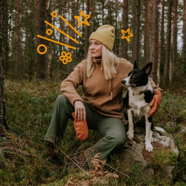 Frau mit blonden Haaren sitzt im Wald auf einem Stein mit ihrem Hund im Arm
